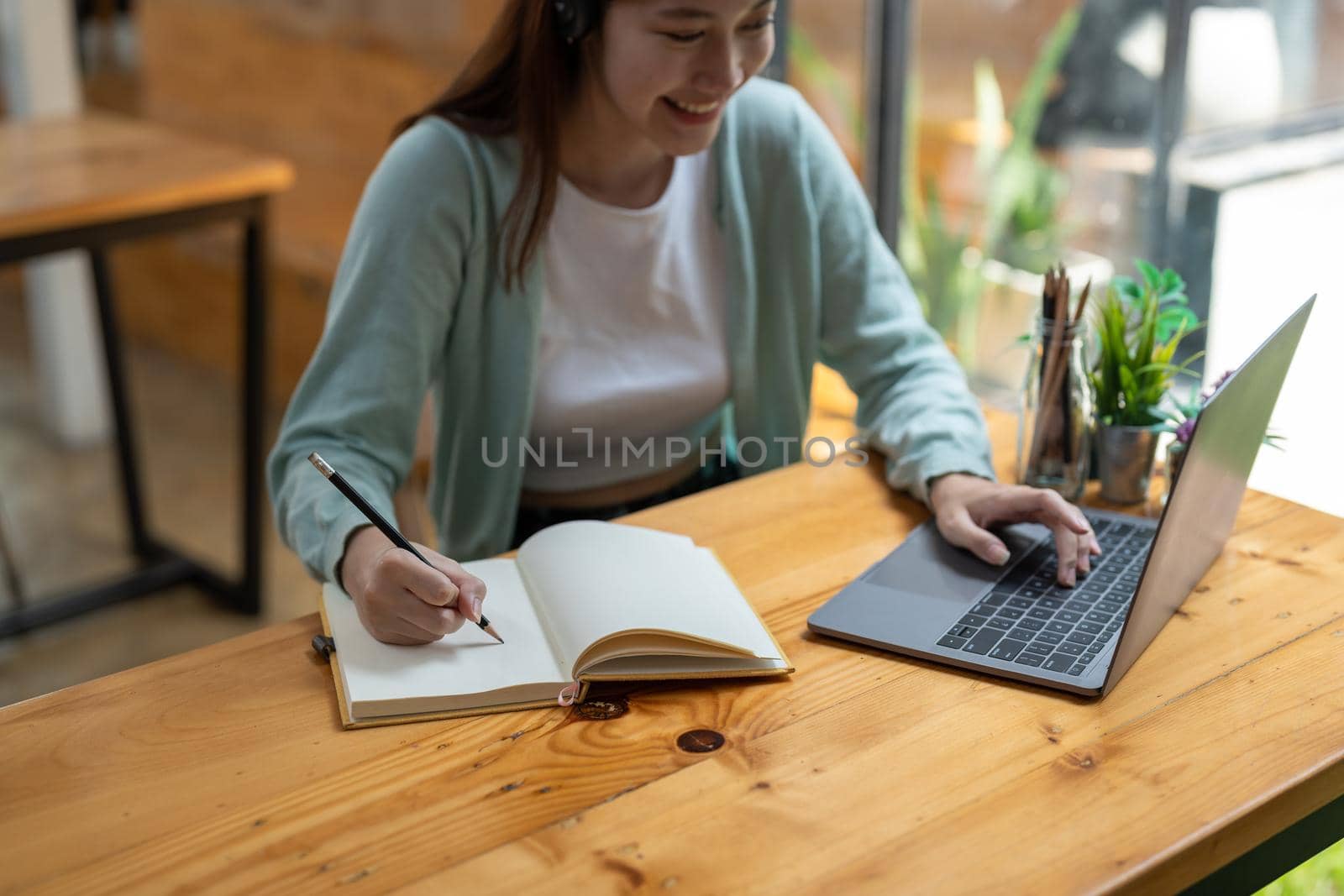 Young Asian girl student wears headphones writing on the notebook to study language online watch and listen to the lecturer, webinar via video call e-learning at home.