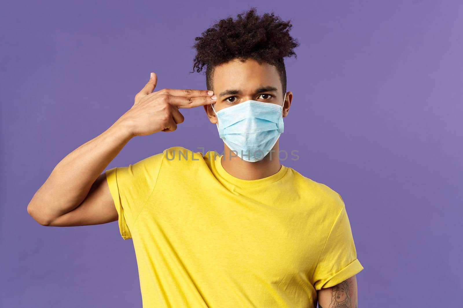 Covid19, healtcare and medicine concept. Close-up portrait of serious hispanic guy in medical mask, showing gun near head as if sick and tired of people not avoid social contact, purple background.