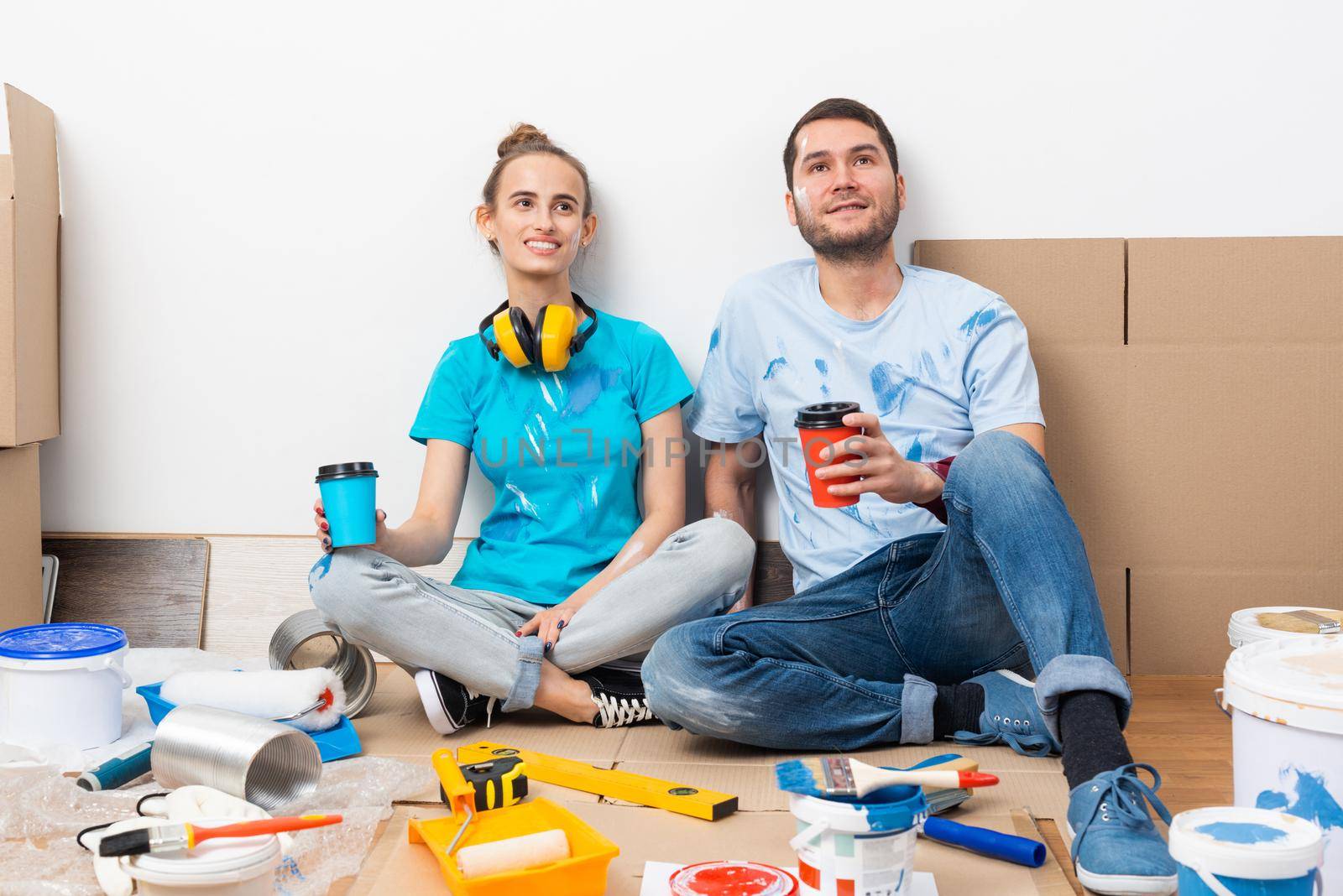 Happy boy and girl drinking coffee on floor. Home remodeling after moving. Cardboard boxes, construction tools and materials for building on floor. Couple having fun in time of house renovation.