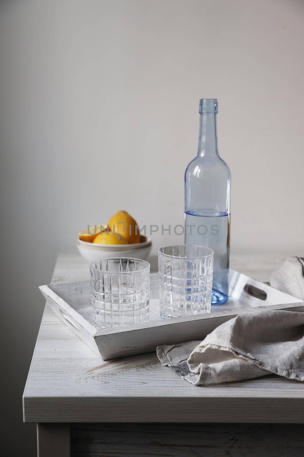 A blue transparent water bottle and two ribbed glasses on a white wooden tray on the kitchen table. by elenarostunova