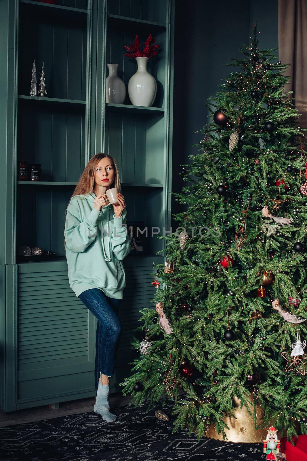 A beautiful woman in homemade clothes near the Christmas tree at home drinks delicious tea or coffee. Festive morning. by StudioLucky