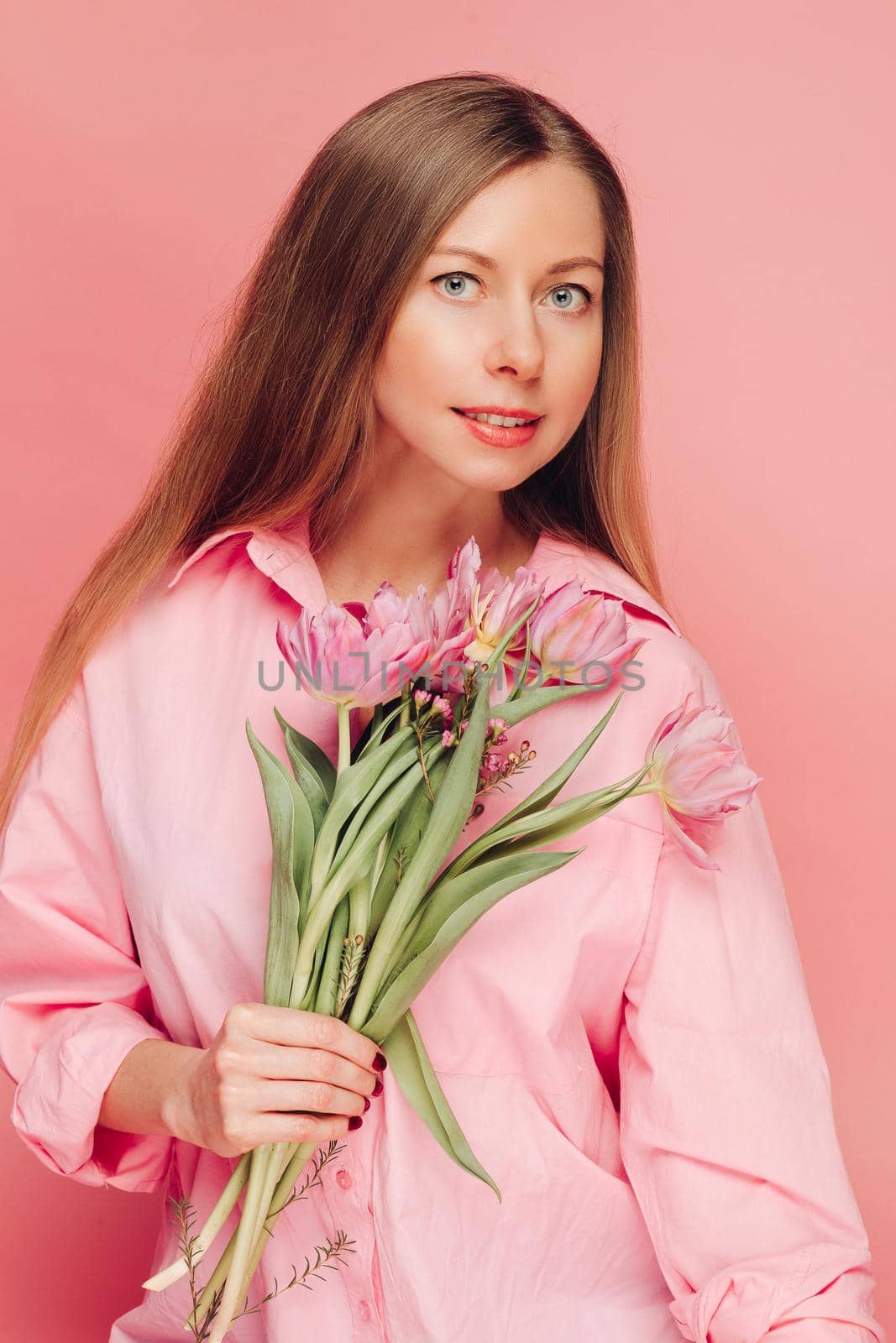 A sweet charming woman with flowers in a pink dress on a pink background smiles, happiness and luck.