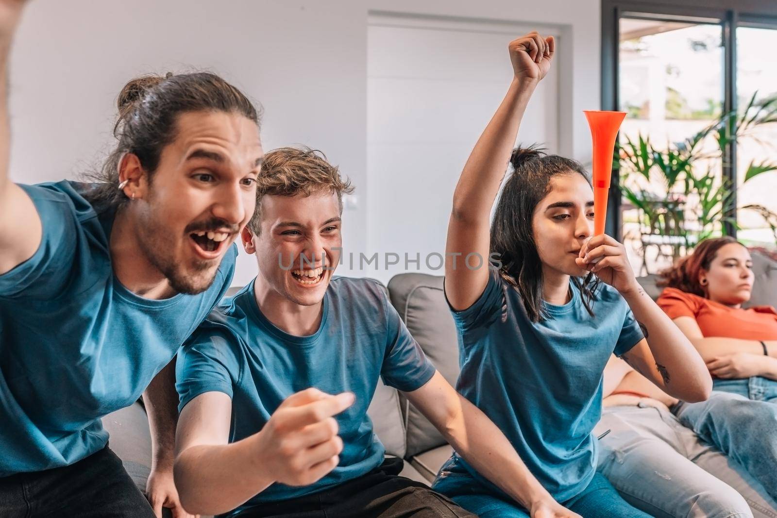 football friends celebrating a victory at home. young people watching sport on TV. leisure concept, three young adults in blue t-shirts. happy and cheerful. natural light in the living room at home.