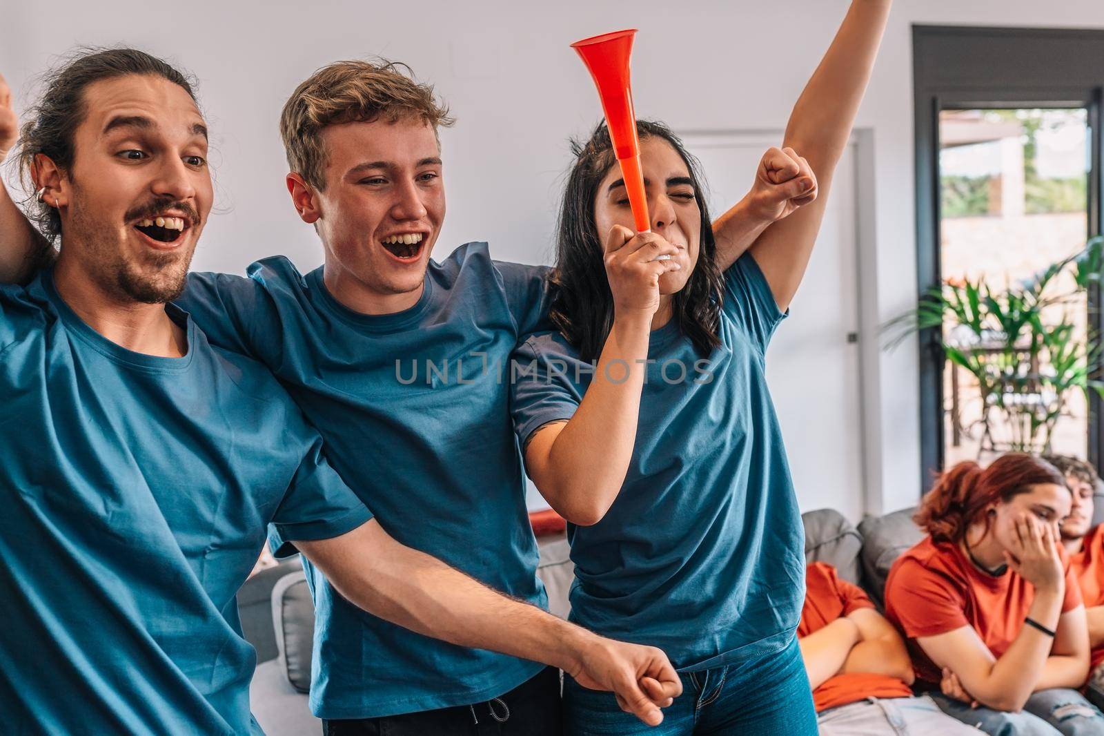 football fan group of friends celebrating a victory at home. young people watching sport on television. leisure concept. three young adults in blue jerseys and red jerseys. happy and cheerful. natural light in living room at home.