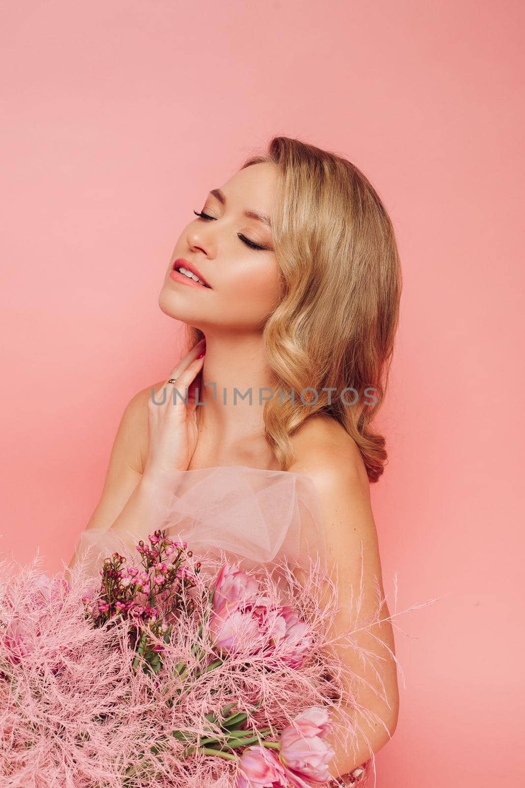 A sweet charming woman with flowers in a pink dress on a pink background smiles, happiness and luck.