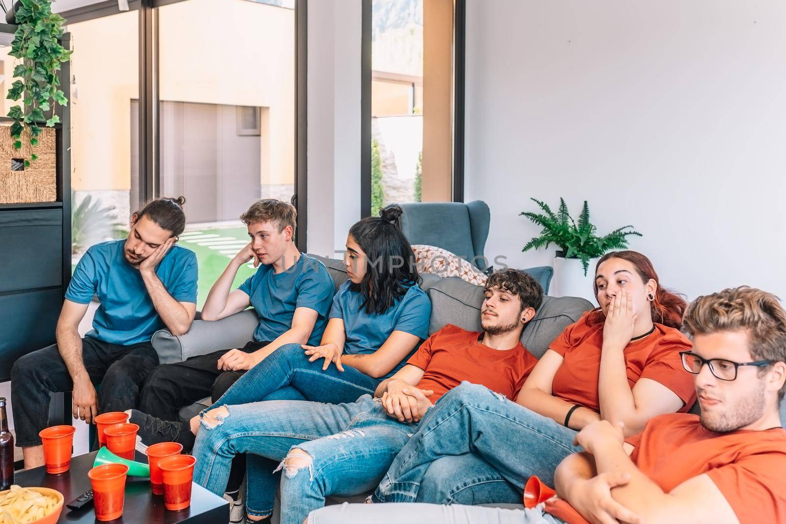 Football fans saddened by their team's defeat. Friends watching football in living room concept of leisure. concept of friendship. group of six people, friendly relationship, divided teams, red and blue, competitive.