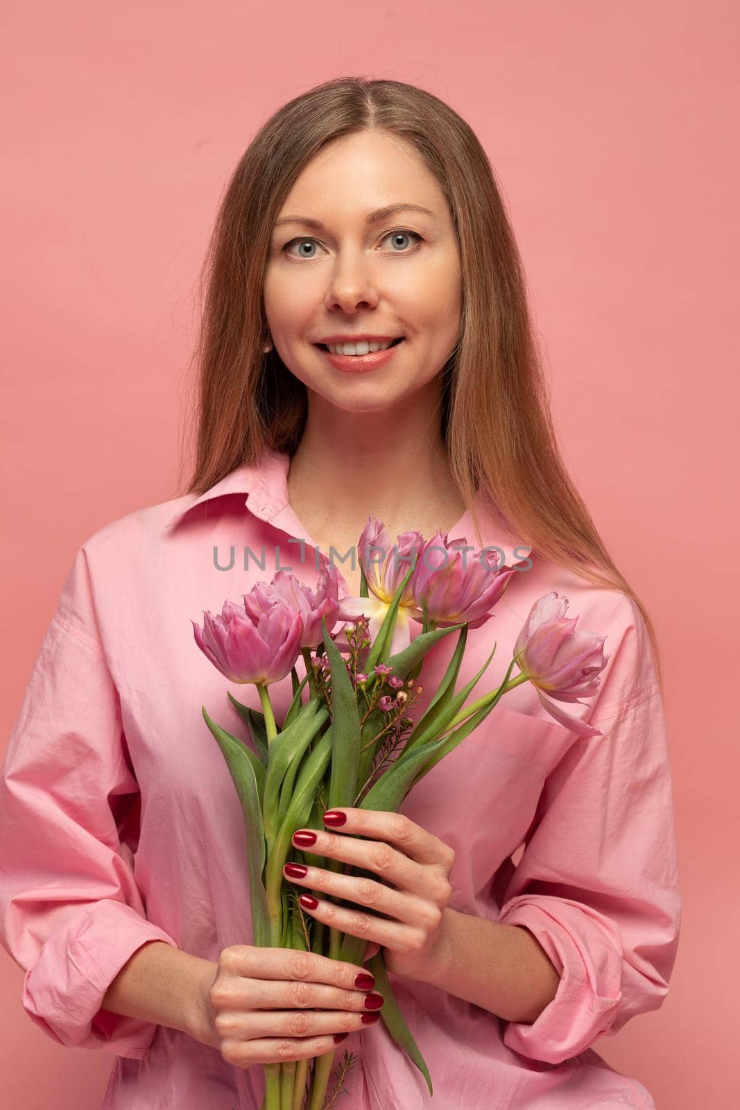 A sweet charming woman with flowers in a pink dress on a pink background smiles, happiness and luck.