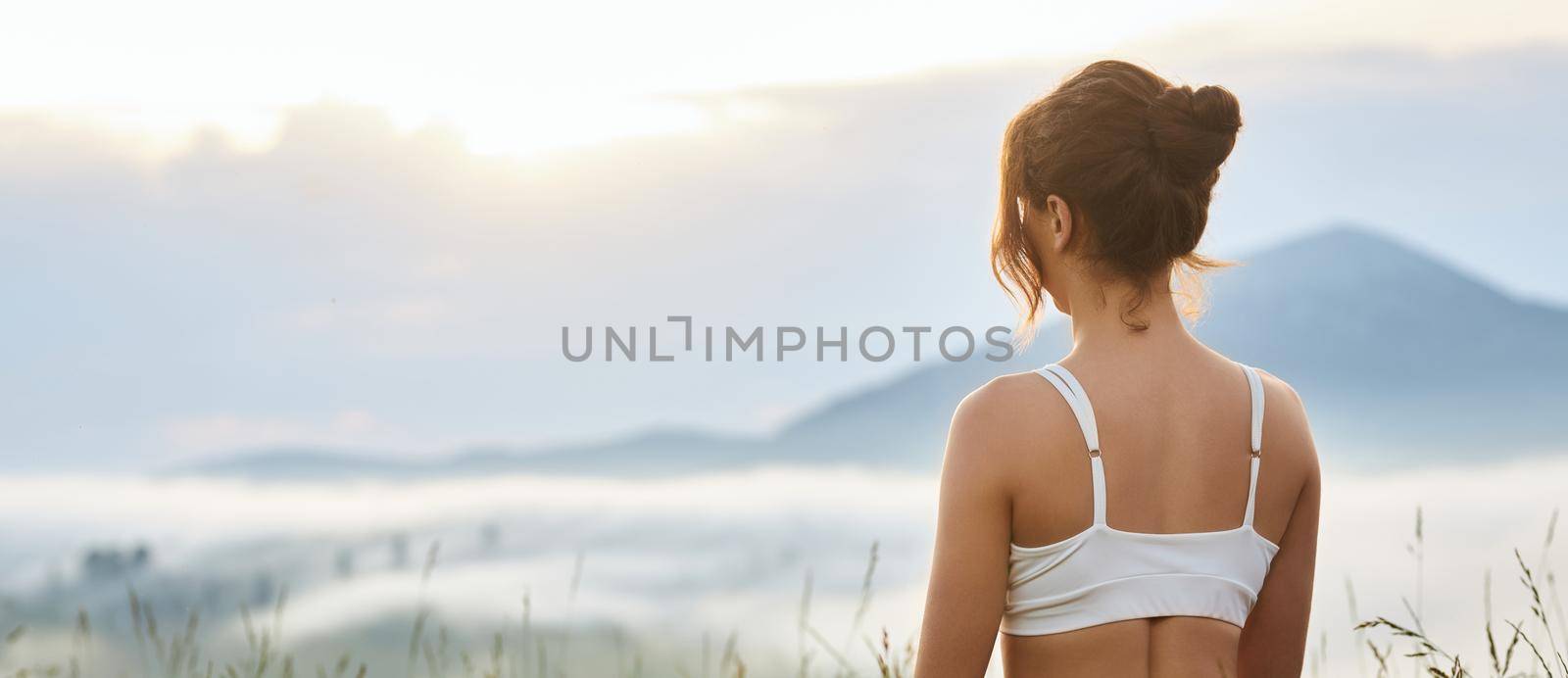 Back panoramic view of woman looking forward in hills. Female with hair bun, wearing white top, enjoying beautiful landscape, dreaming. Concept of harmony with nature.