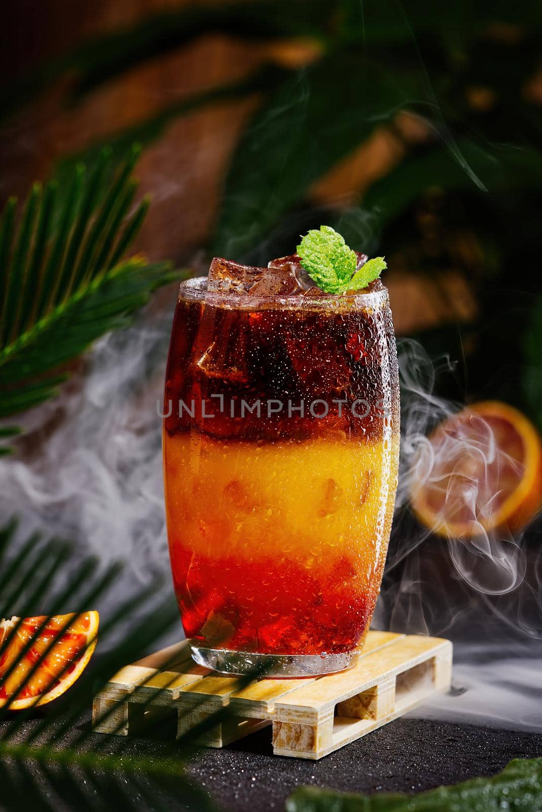 Bumble coffee with ice on a dark tropical background. Espresso, orange juice and syrup in layers in transparent glass with ice. Summer trendy refreshing coffee drink. Coffee house menu, recipe. Copy space