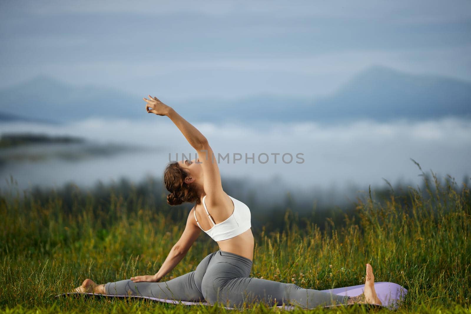 Side panoramic view of female practicing yoga in hills. Pretty girl sitting on twine on green grass alone with raised hand in nature. Concept of new age.