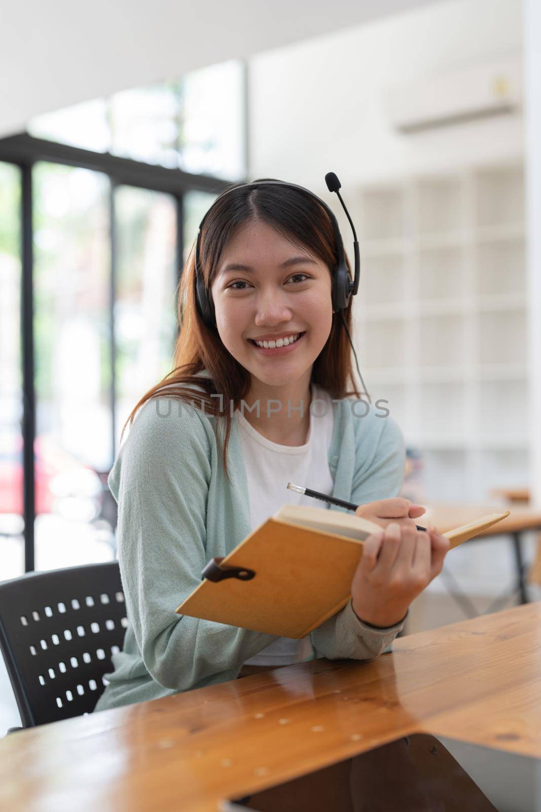 Portrait young asian woman with headphone writing making list taking notes in notepad working or learning on laptop indoors- educational course or training, seminar, education online concept by nateemee