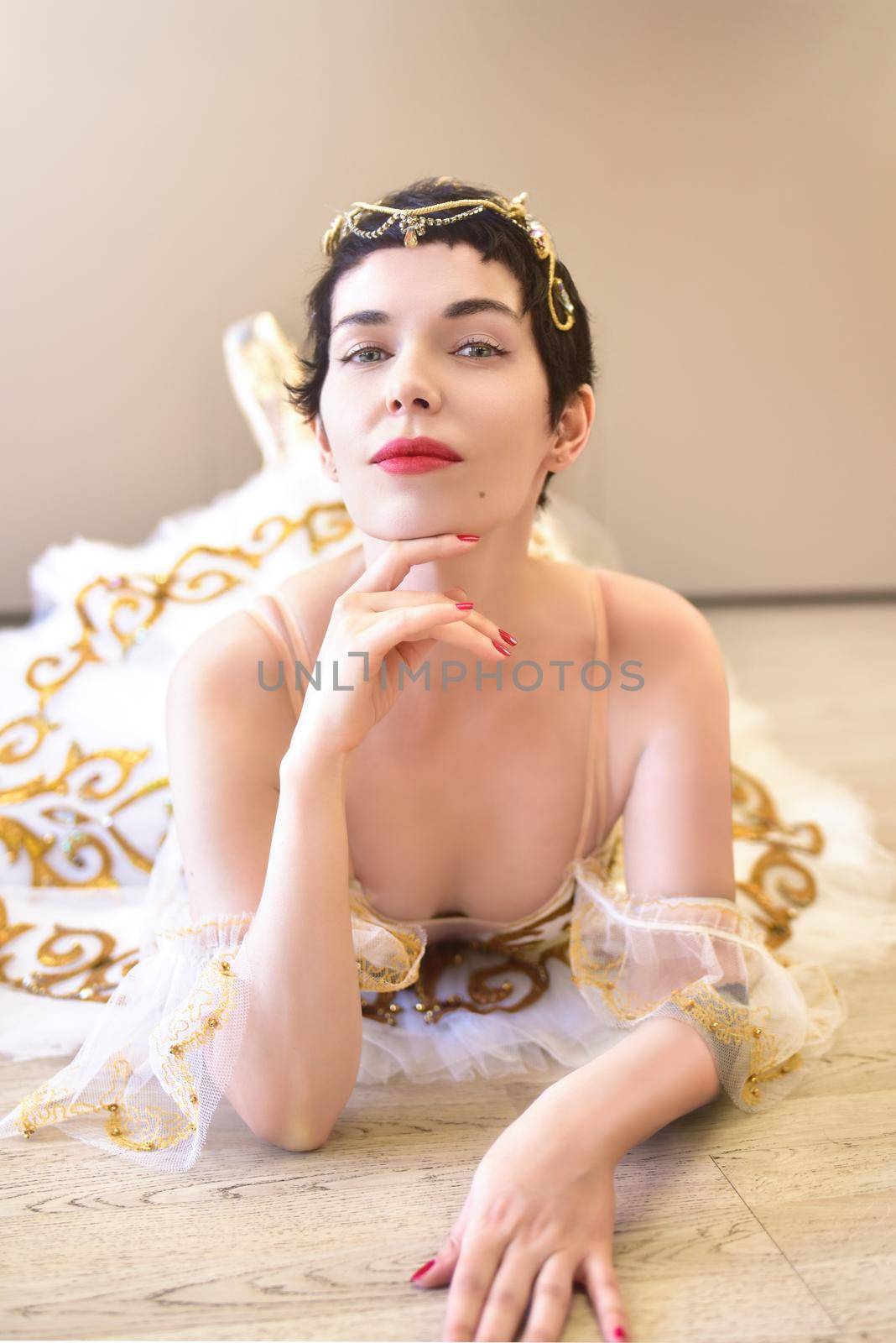 A portrait of a charming ballerina lying on the floor in a ballroom, she touches her chin with her hand and looks flirtatiously at the camera