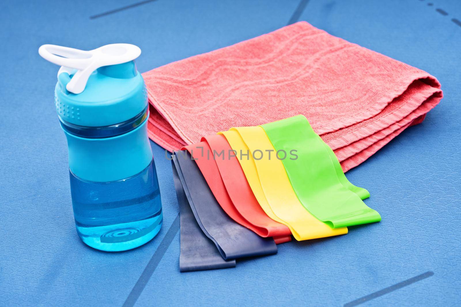 Close up of yoga or fitness items – elastic resistance rubber bands, towel, and water bottle on a blue mat. Sports Concept. Sports accessories.