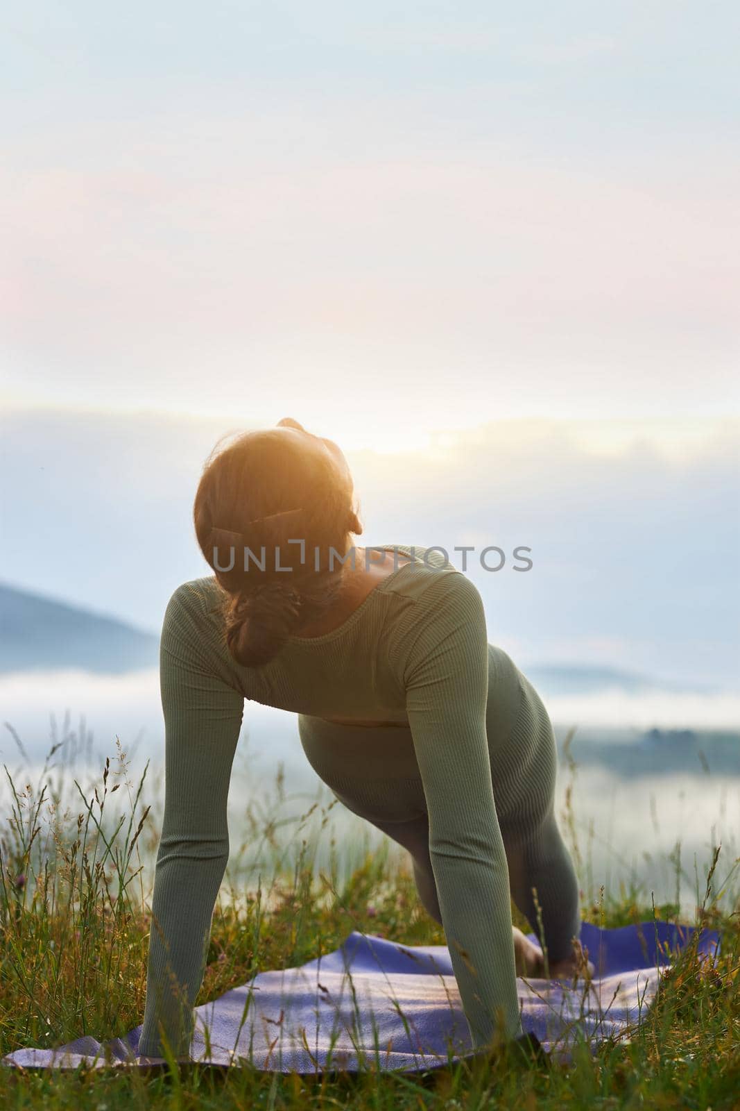 Back view of young woman practicing yoga position in mountains. Flexible and slim female standing in plank on yoga mat wearing sport suit in hills. Concept of yoga.