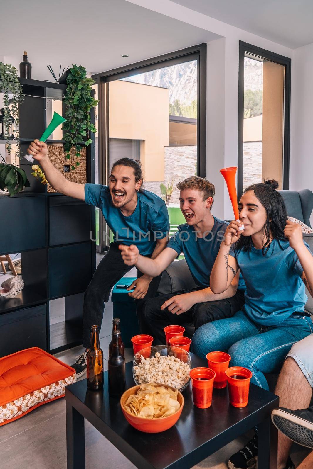 friends of championship fans shouting for joy after their team's victory, watching the broadcast on television. leisure concept, three young adults in blue t-shirts. happy and cheerful. natural light in the living room at home. trumpet