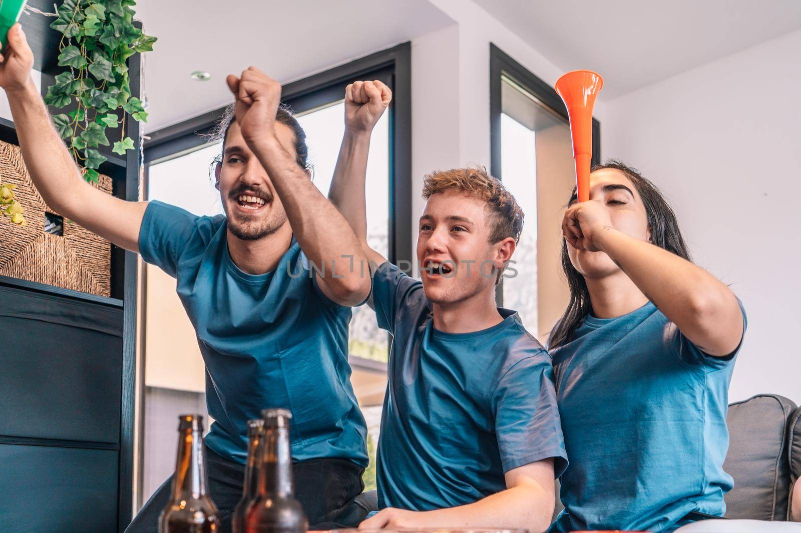 football fan group of friends celebrating a victory at home. young people watching sport on television. leisure concept. three young adults in blue jerseys and red jerseys. happy and cheerful. natural light in living room at home.