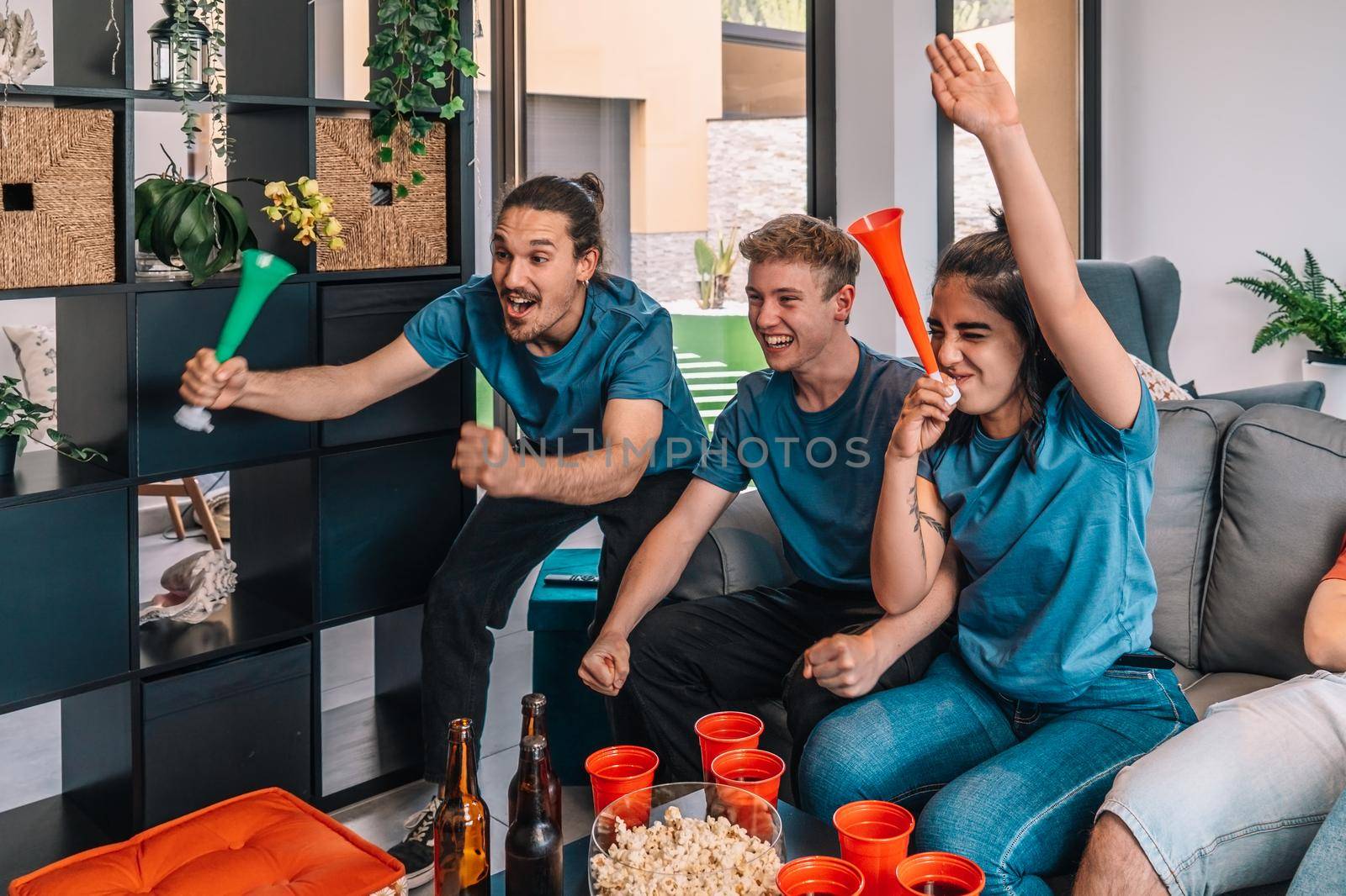 friends of championship fans shouting for joy after their team's victory, watching the broadcast on television. leisure concept, three young adults in blue t-shirts. happy and cheerful. natural light in the living room at home. trumpet