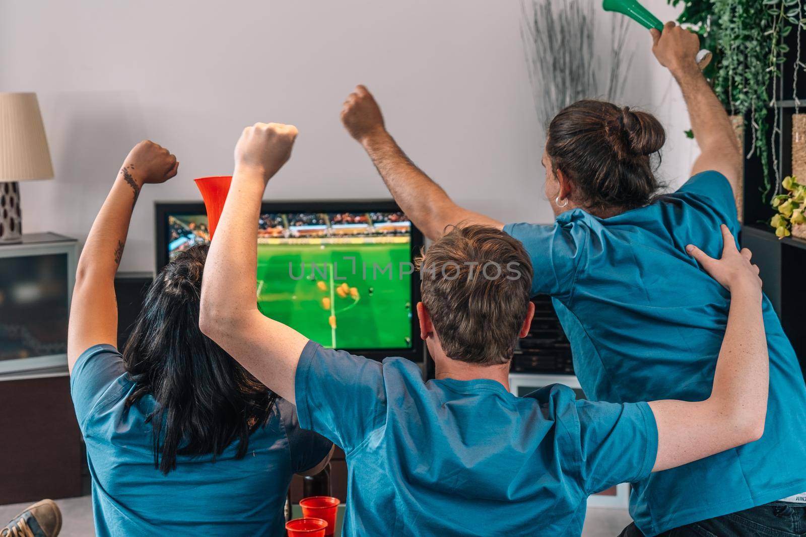 football friends celebrating a victory at home. young people watching sport on TV. leisure concept, three young adults in blue t-shirts. happy and cheerful. natural light in the living room at home.