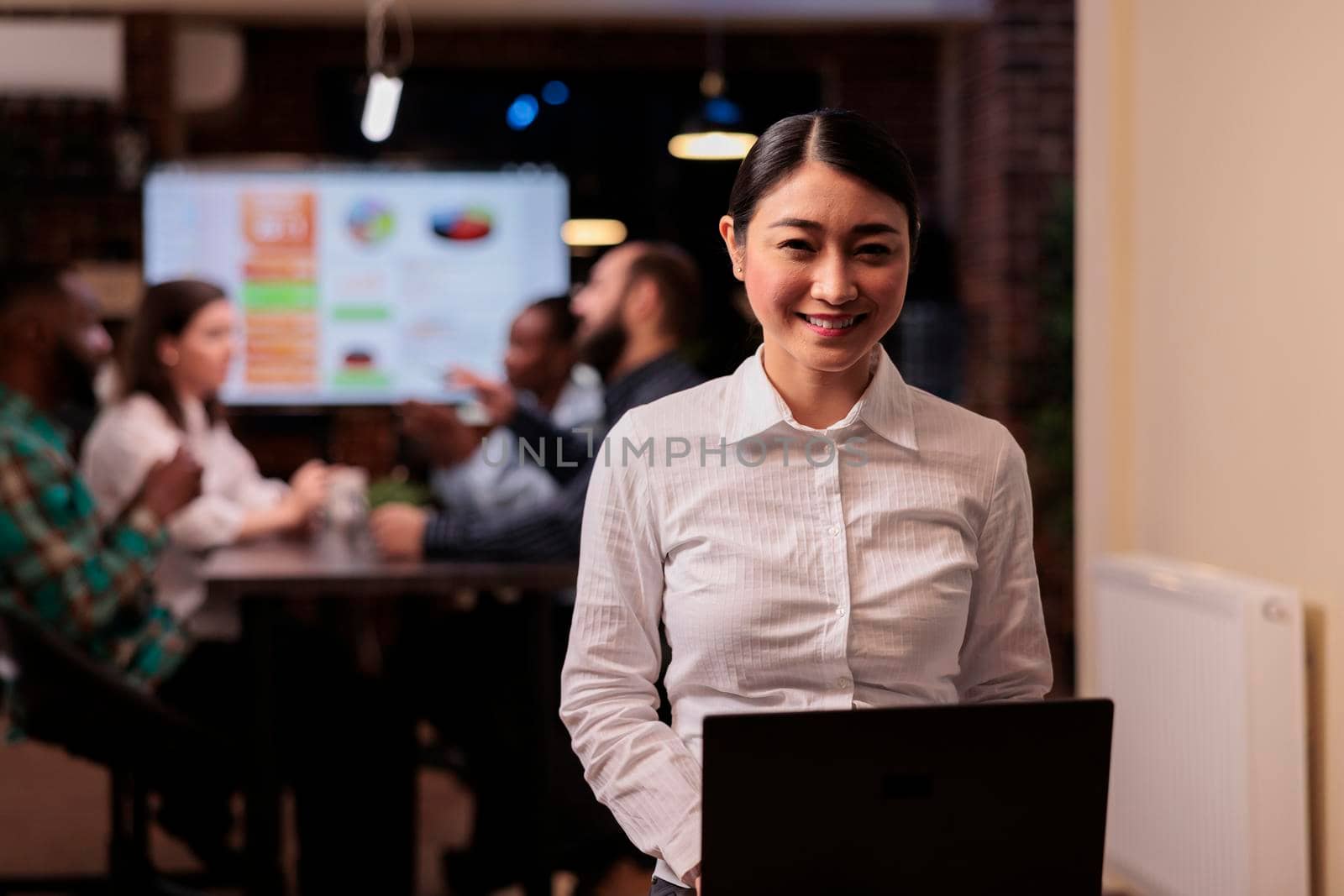 Portrait of smiling asian business employee holding laptop in startup office working overtime by DCStudio