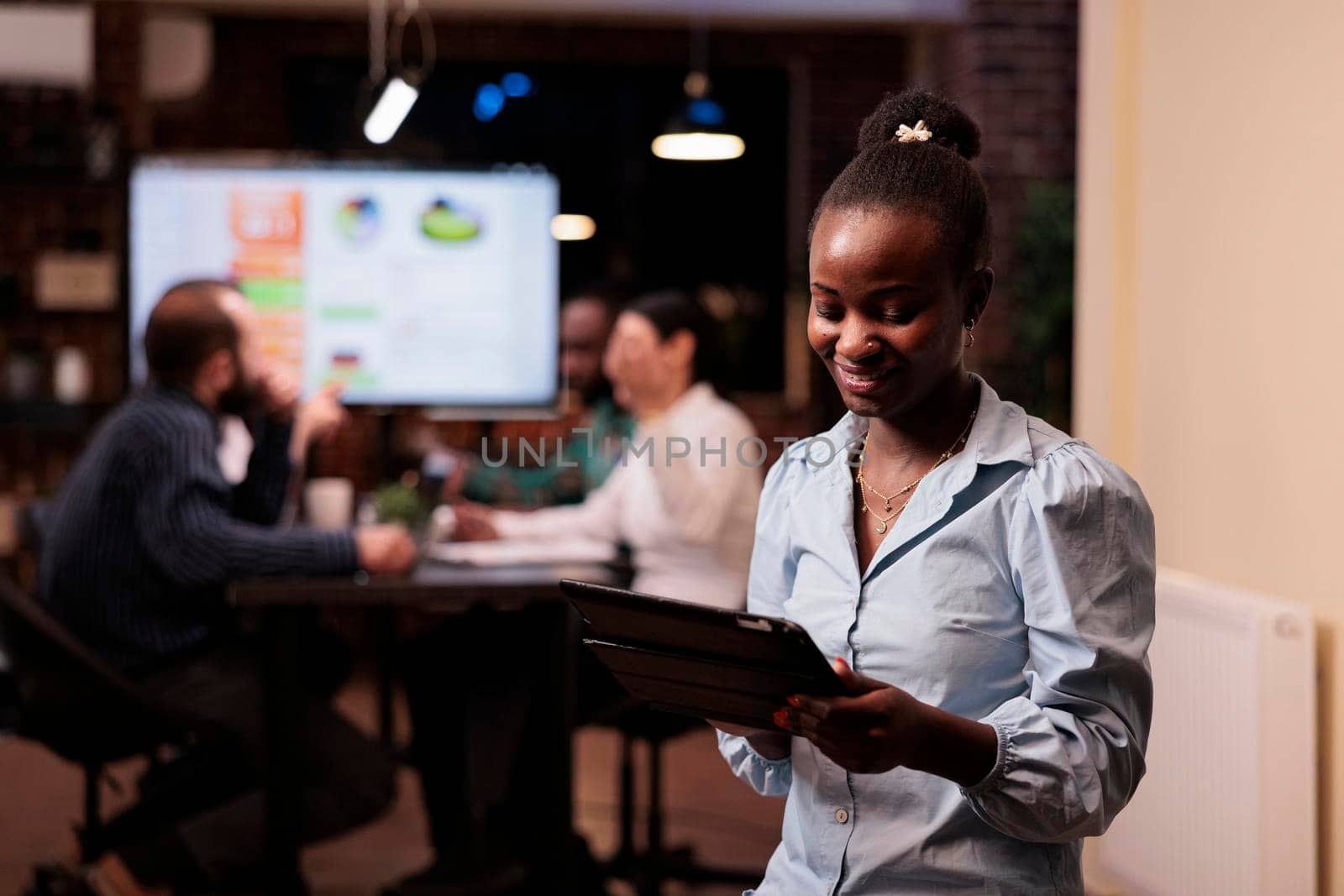 Portrait of african american woman holding digital tablet and looking at screen working late hours with mixed team. Startup employee posing confident using touchscreen device in group project.