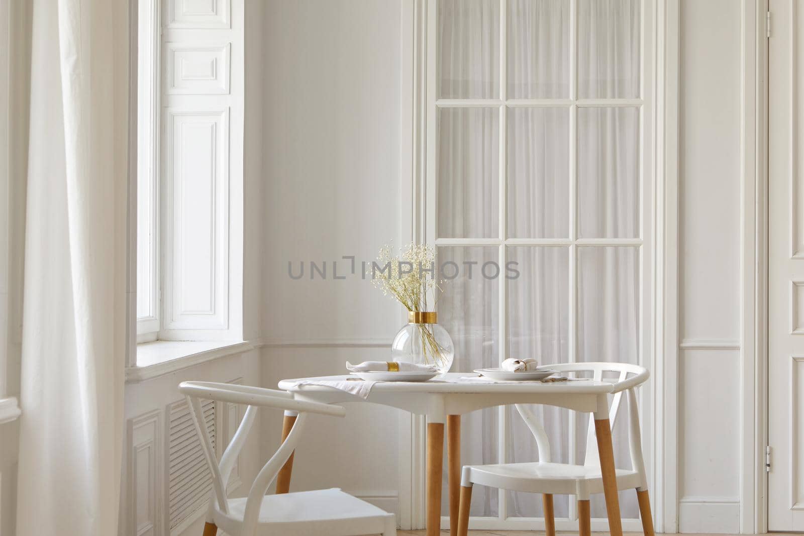 Glass vase with dried flowers and dishware placed on table near chairs and window in white spacious room at home