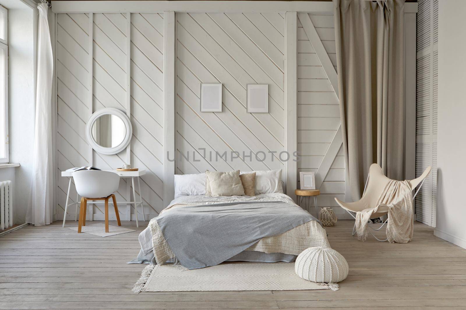 Comfortable bed with blanket and cushions placed on rug at wall with decorative frames in stylish bedroom with table and armchair in pastel colors