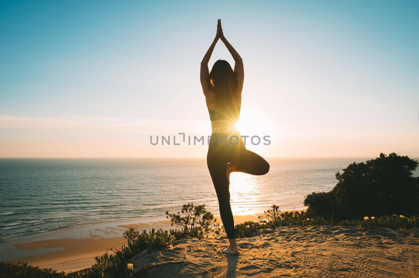 Woman practicing yoga and meditates on the mountain with ocean view at sunset. Young woman standing in vrksasana exercise, tree pose outdoor in the nature. High quality photo