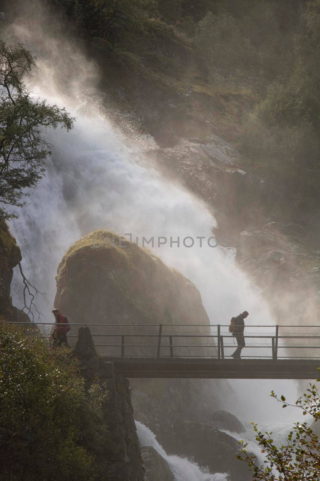 Misty landscape showing men walking on a bridge and a big waterfall falling over a rock behind them
