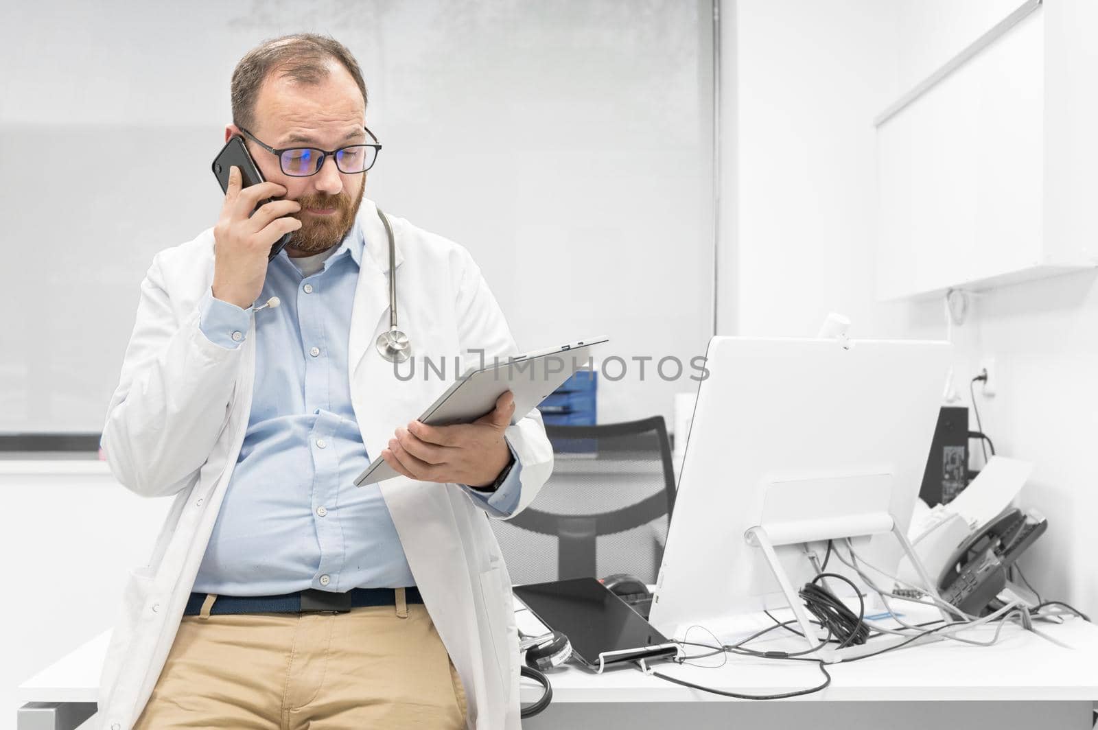Male physician with stethoscope sitting at desk, talking on mobile phone and looking at digital tablet, discussing a patient diagnostic. by HERRAEZ