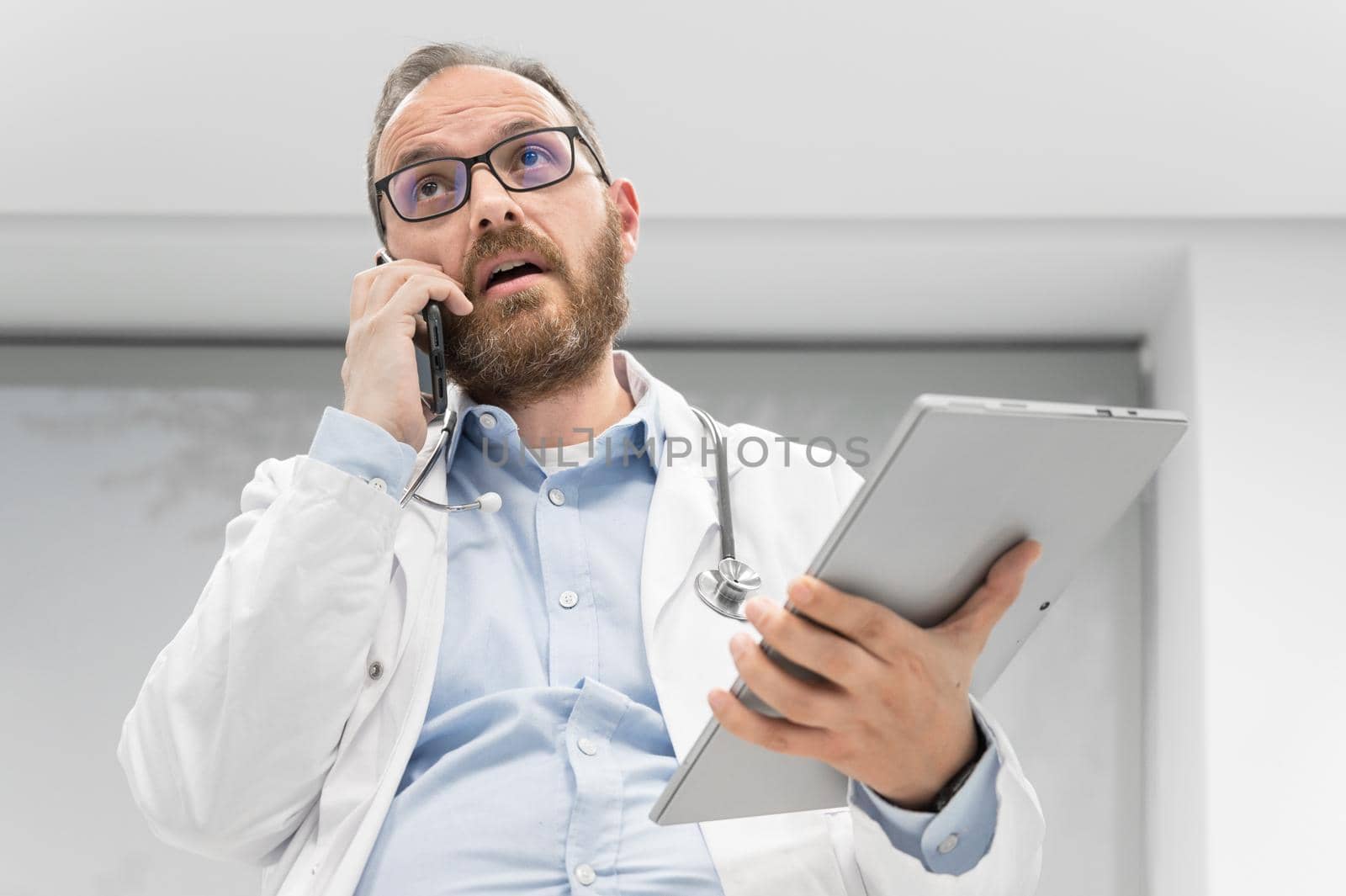 Male physician with stethoscope sitting at desk, talking on mobile phone and looking at digital tablet, discussing a patient diagnostic. by HERRAEZ