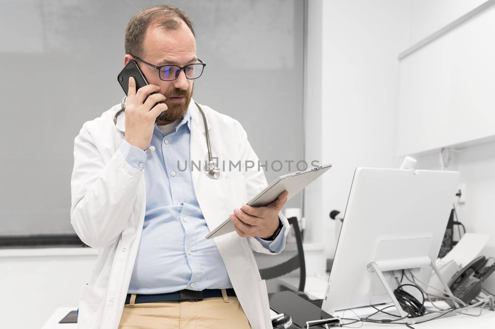 Male physician with stethoscope sitting at desk, talking on mobile phone and looking at digital tablet, discussing a patient diagnostic. High quality photography