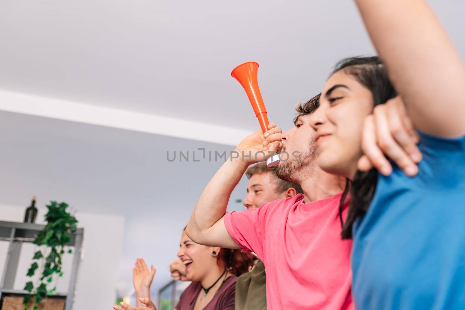 friends cheering for their team after scoring a point in their favour. sport broadcast on television. group of young people partying at home. leisure concept. three young adults in blue jerseys and red jerseys. happy and cheerful. natural light in living room at home.