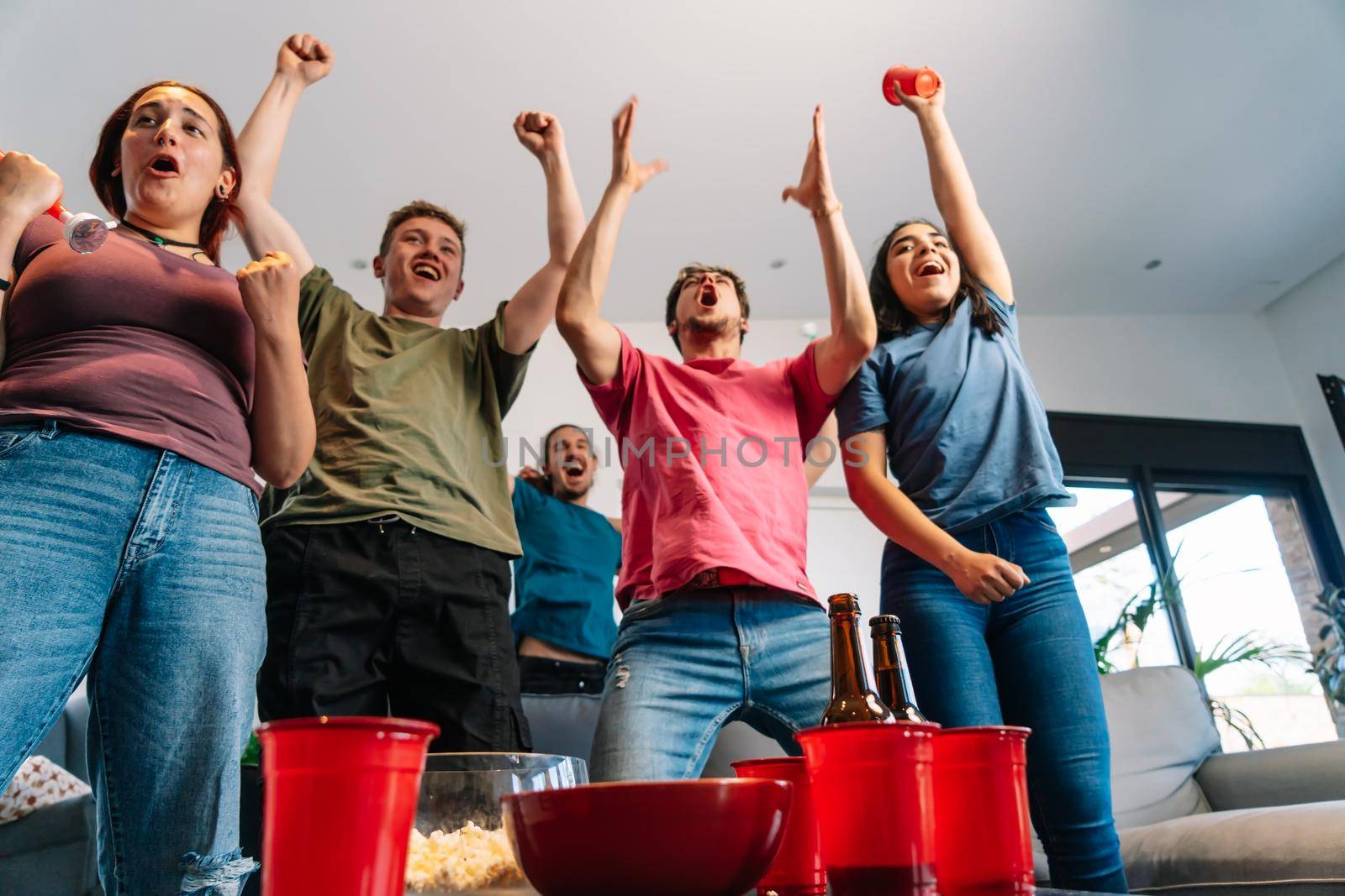 friends jumping for joy watching e-sports on TV after their team's victory. group of young people partying at home. leisure concept. happy and cheerful. natural light in the living room at home. trumpet