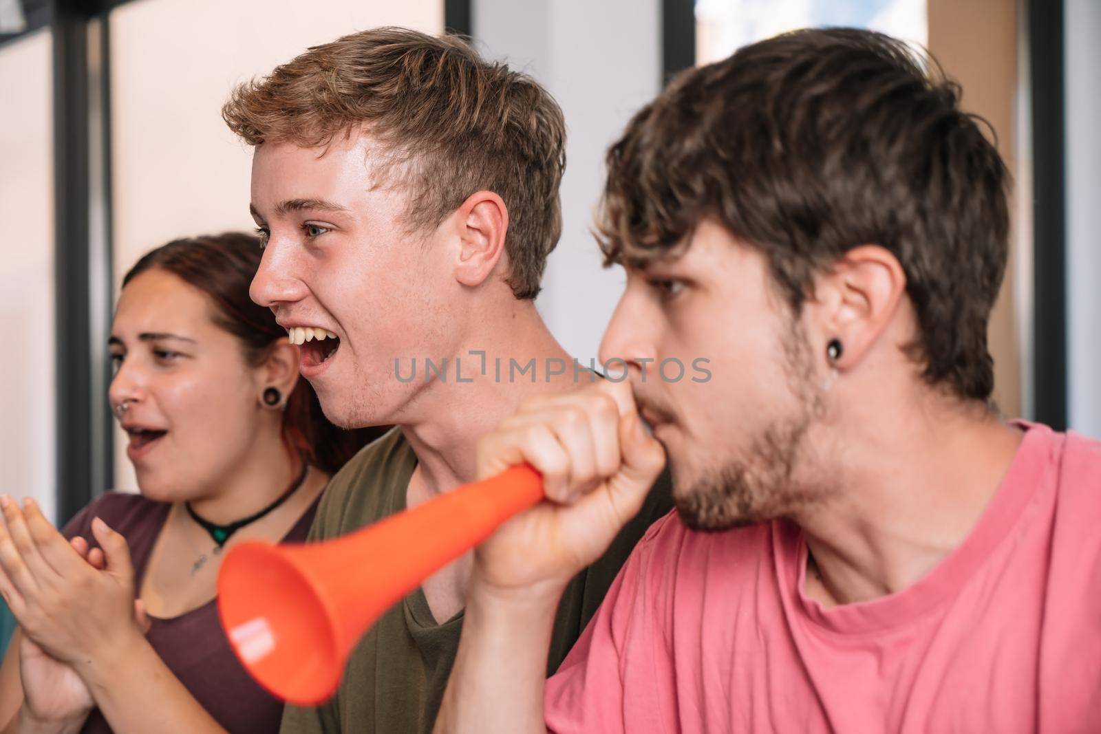 football fan group of friends celebrating a victory at home. young people watching sport on television. leisure concept. three young adults in blue jerseys and red jerseys. happy and cheerful. natural light in living room at home.