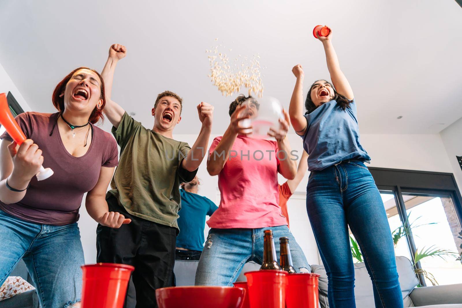 friends jumping for joy, throwing popcorn in the air after their team's victory. group of youngsters partying at home. by CatPhotography