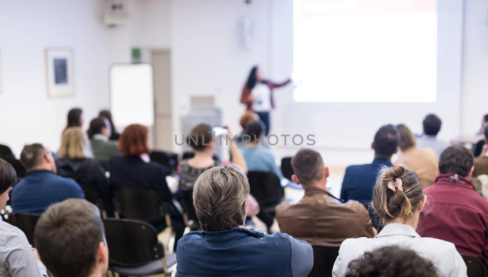 Business and entrepreneurship symposium. Female speaker giving a talk at business meeting. Audience in conference hall. Rear view of unrecognized participant in audience. Copy space on whitescreen.