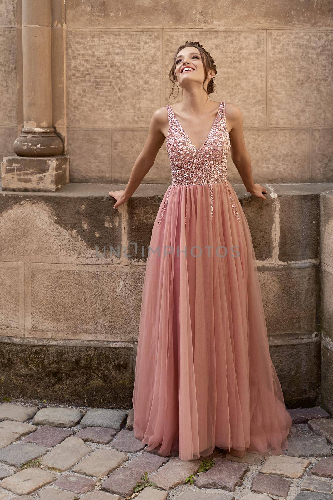 Beautiful brunette bridesmaid with luxury hairdo in gorgeous elegant stylish pale pink chiffon gown dress on flowers background. Wedding day in old beautiful European city.