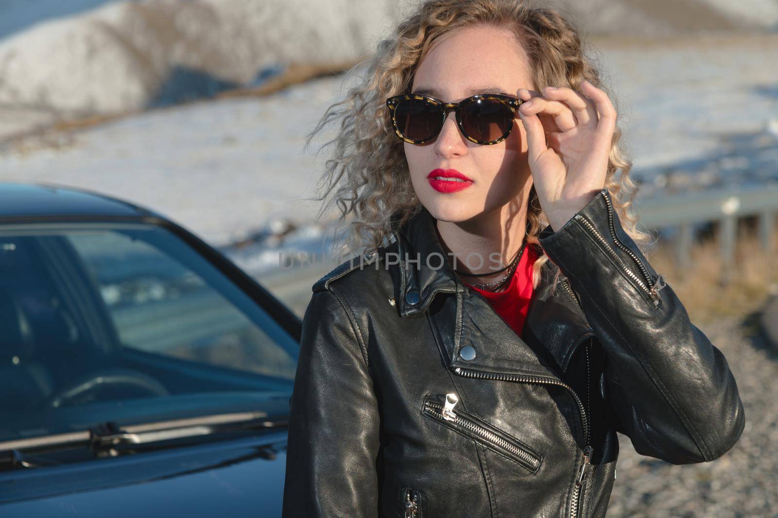 Street fashion portrait of young elegant luxury lady in sunglasses, curly hair and leather jacket posing near retro car. Copy, empty space for text. Holding on to sunglasses in the mountains.