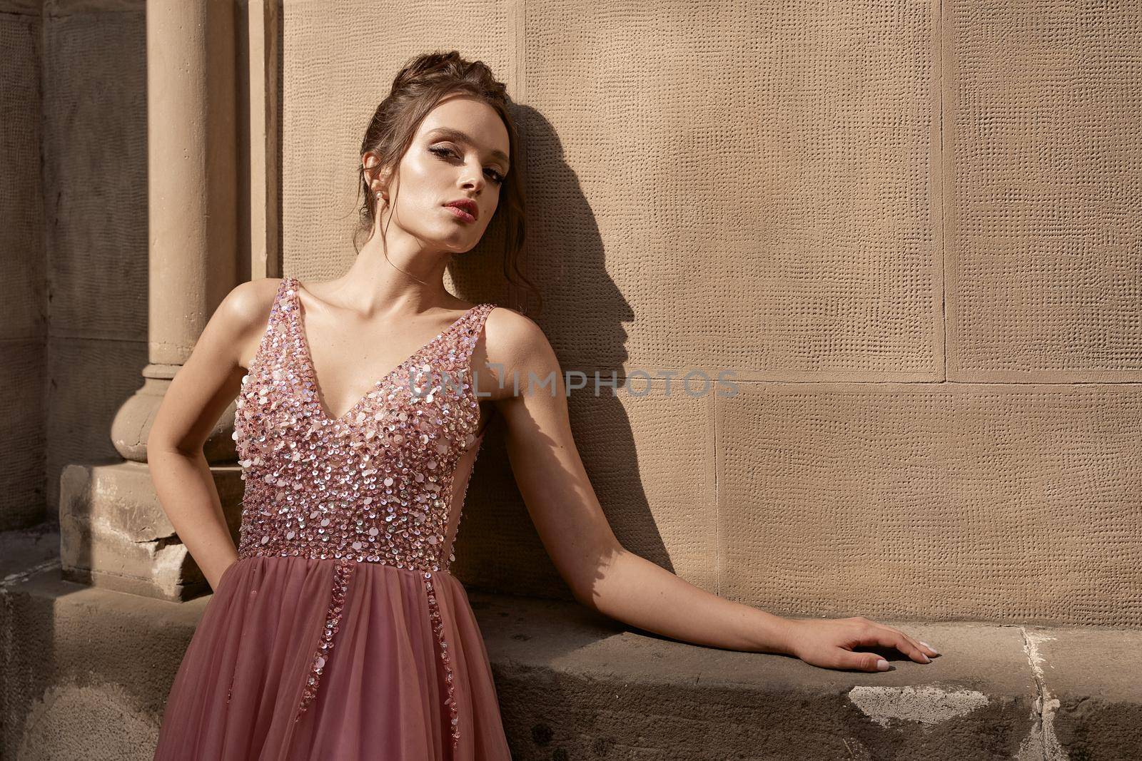 Beautiful brunette bridesmaid with luxury hairdo in gorgeous elegant stylish pale pink chiffon gown dress on flowers background. Wedding day in old beautiful European city.