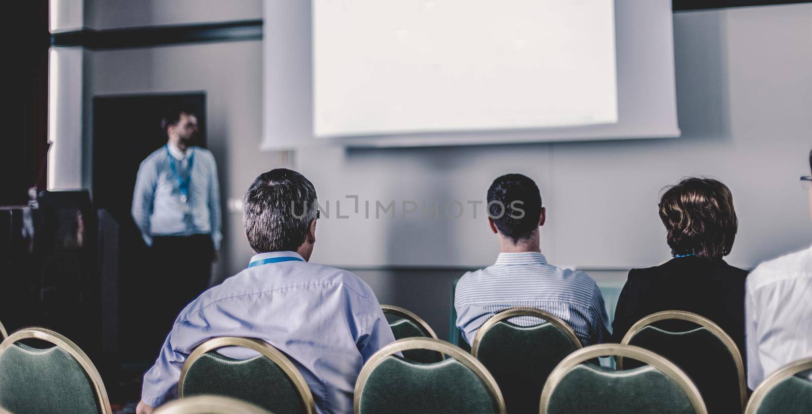 Business and entrepreneurship symposium. Speaker giving a talk at business meeting. Audience in the conference hall. Rear view of unrecognized participant in audience.