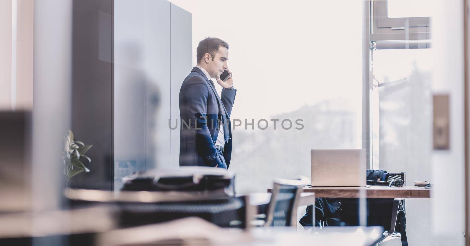 Businessman talking on a mobile phone while looking through modern corporate office window.