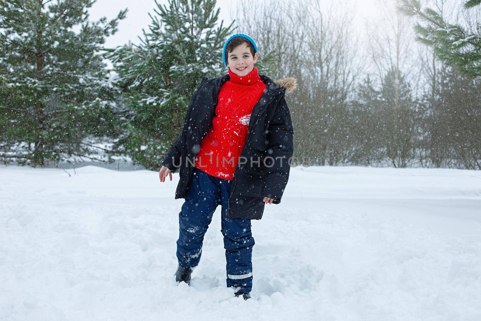 A cute teenager in a dark jacket and a red sweater stands in a winter park on the snow