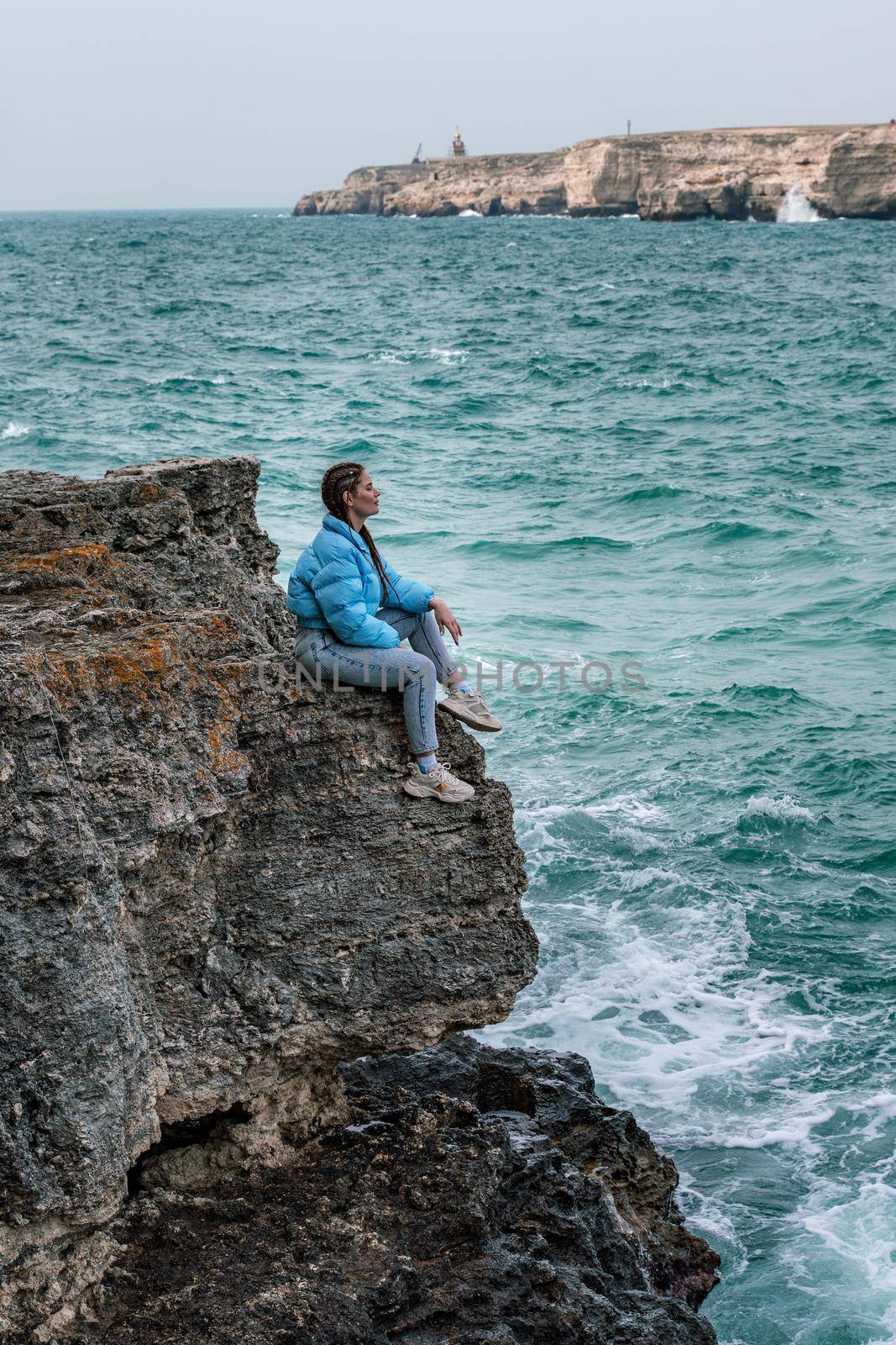 A woman in a blue jacket sits on a rock above a cliff above the sea, looking at the stormy ocean. Girl traveler rests, thinks, dreams, enjoys nature. Peace and calm landscape, windy weather. by Matiunina