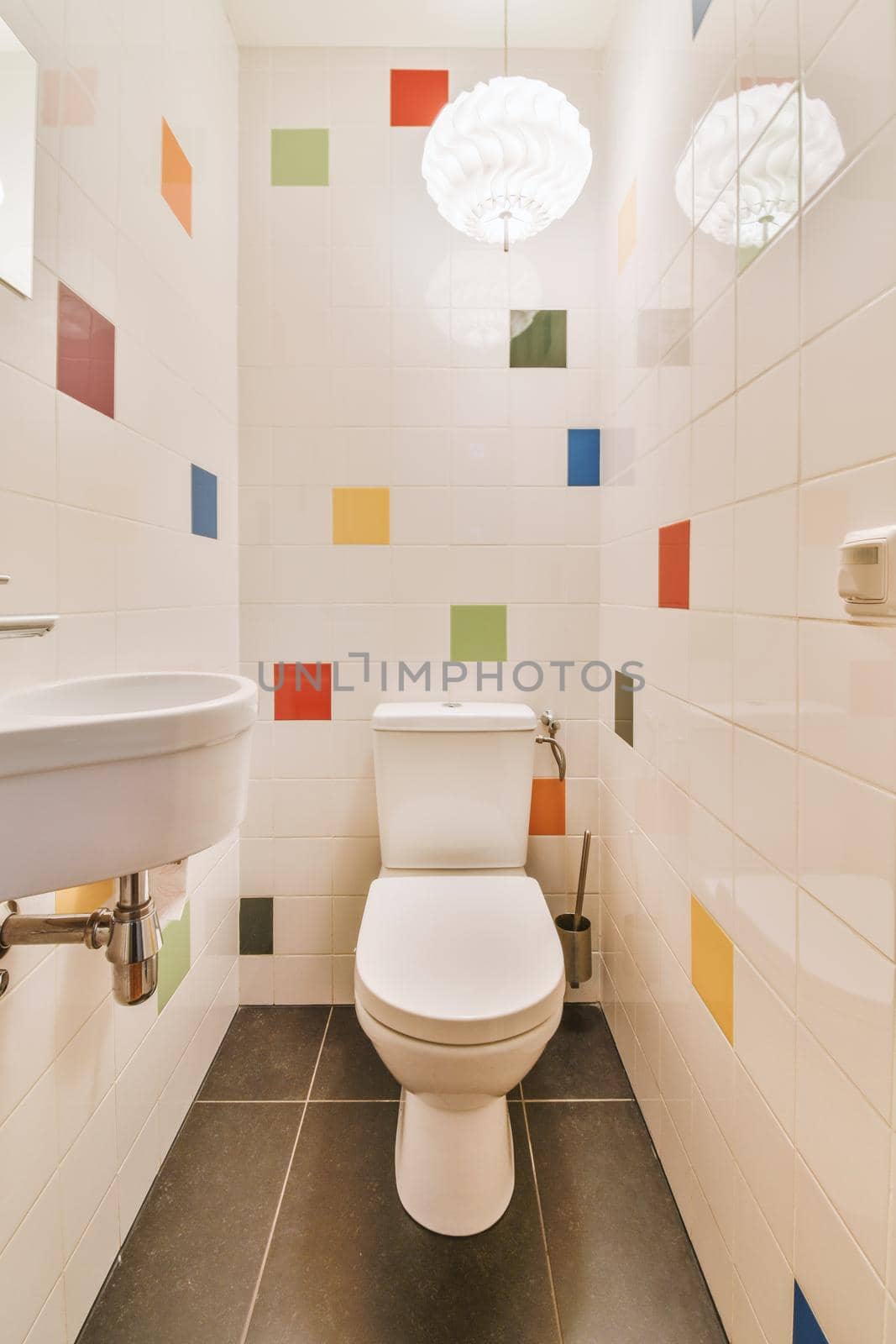Toilet and small sink in narrow lavatory room with colourful tile and bright lamp