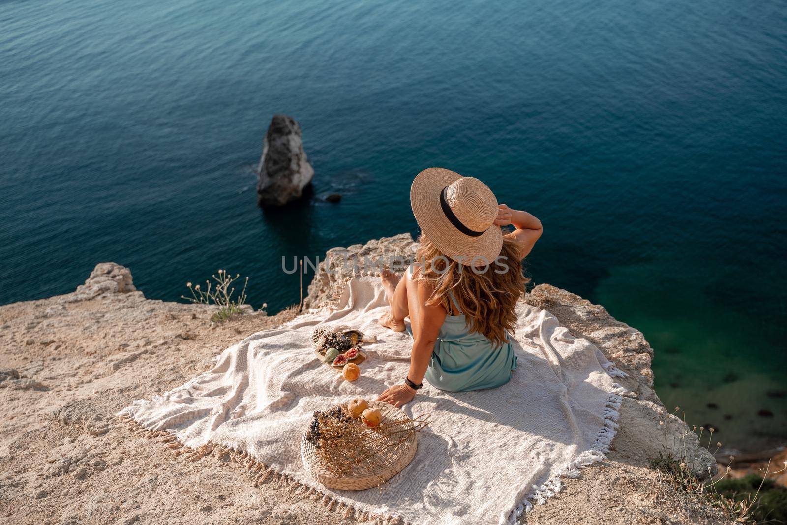 Street photo of a beautiful woman with dark hair in a mint dress and hat having a picnic on a hill overlooking the sea. by Matiunina