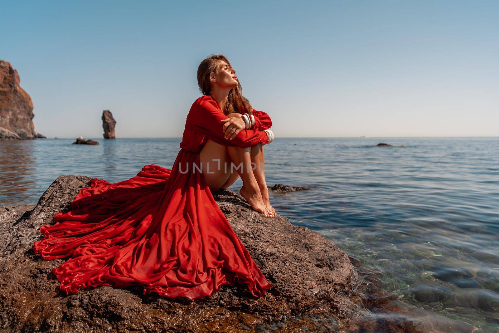 Beautiful sensual woman in a flying red dress and long hair, sitting on a rock above the beautiful sea in a large bay. by Matiunina