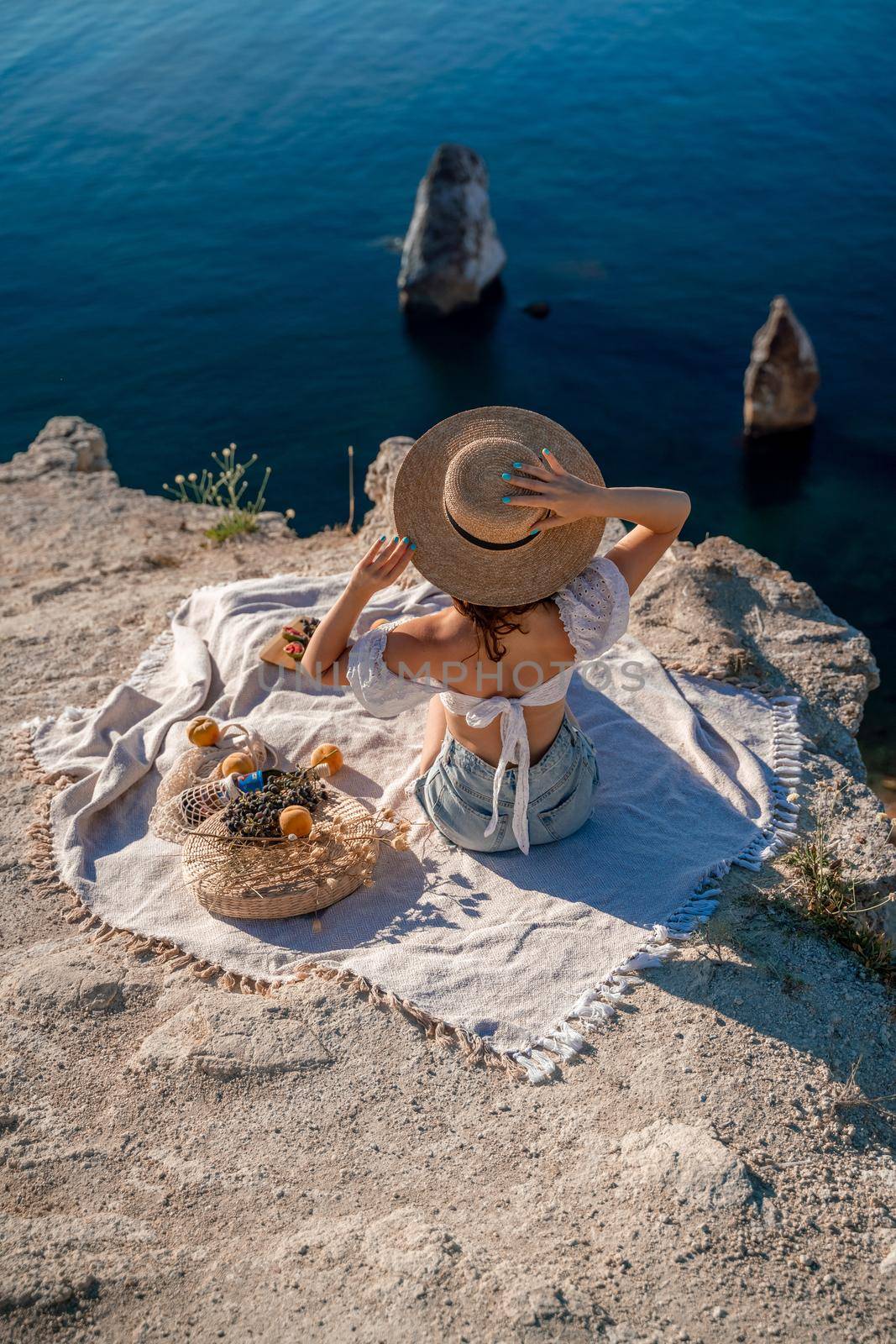 Street photo of a beautiful woman with dark hair in a white top, shorts and a hat having a picnic on a hill overlooking the sea.