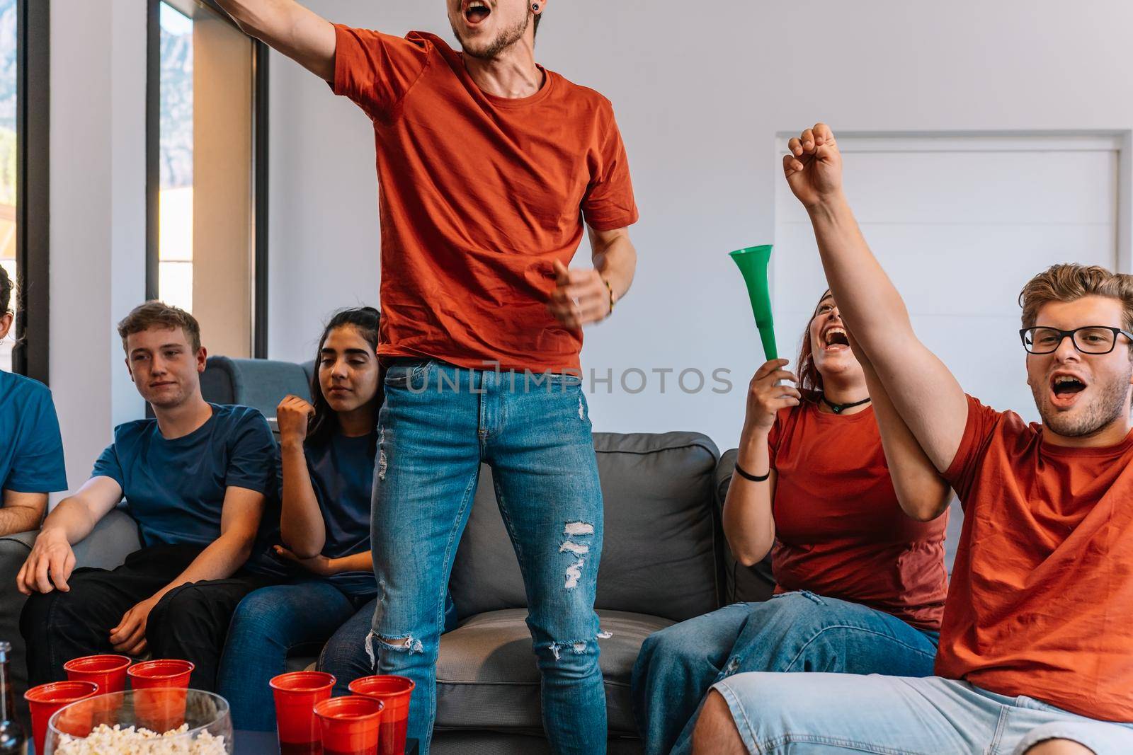 friends jumping for joy after their team's victory, watching football match on TV. opposing team sad after defeat leisure concept, three young adults in blue t-shirts. happy and cheerful. natural light in the living room at home.