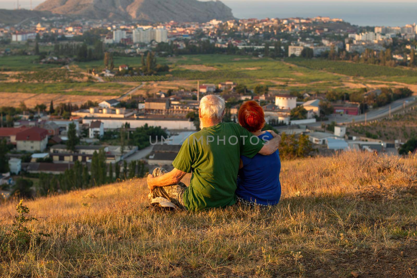 An elderly couple sits on a mountain with their backs with a beautiful view of the mountains and the sea in the distance