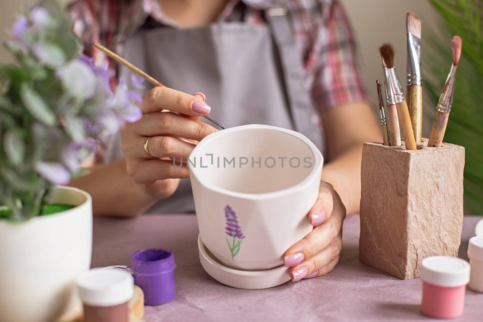 A hands of woman in a plaid shirt and a gray apron paints a white ceramic flower pot at the table. On the table is purple kraft paper, brushes, jars of paints. Close-up. No face